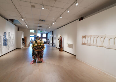 A contemporary art gallery showcasing sculptures and textile artworks crafted with sustainable art practices. Spot lighting, a wood floor, and a large window at the far end highlight each piece’s unique blend of creativity and sustainability.
