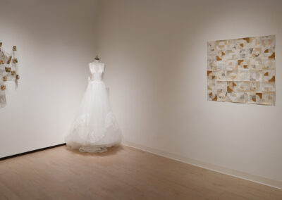 A white bridal gown on a dress form stands in a gallery, showcasing the Art of ReUse. To the left, sheer fabric with small photos is displayed, while to the right, a large patchwork collage of paper pieces highlights sustainable art.