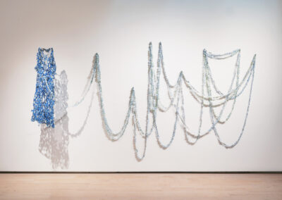 A blue sculptural dress, created through the Art of ReUse, hangs on a gallery wall, its intertwined materials cascading in looping chains of pale blue and white—resembling draped garlands or undulating waves.