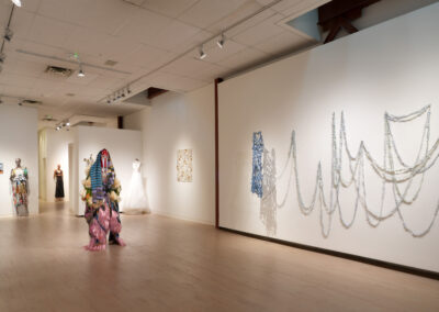 A contemporary art gallery with sculptures, fabric installations on the wall, dresses on mannequins, and a person in a colorful, layered costume—celebrating the Art of ReUse and sustainable art at the center of creative expression.