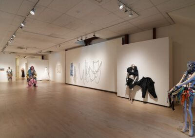 A contemporary art gallery showcasing the Art of ReUse, with mannequins dressed in colorful, eclectic costumes made from reused materials, abstract wall art, and hanging chain installations in a spacious room with wooden floors and white walls.