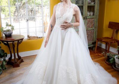 A bride in a white lace wedding gown and veil stands on a floral rug in a bright room with yellow walls, large windows, plants, wooden furniture, and a green antique cabinet—a charming scene inspired by the Art of ReUse.