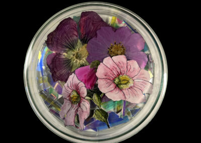 A glass dish containing a colorful arrangement of pressed flowers, including pink and purple blooms, displayed on iridescent shards, showcases the Art of ReUse against a striking black background.