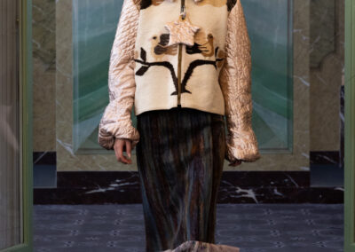 A model wears a cream hooded jacket with animal ear details and a black-brown abstract design inspired by the Art of ReUse, a dark pleated skirt with sheer ruffled hem, black heels, and oval glasses, walking indoors with green marble walls.