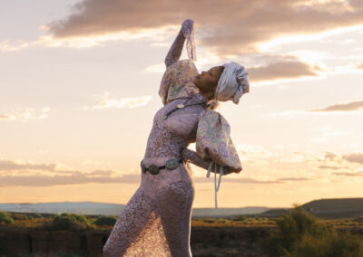 A woman in a long, patterned lavender dress and headwrap poses gracefully outdoors at sunset, arching her back and lifting one arm, embodying the Art of ReUse amid dramatic clouds and a scenic landscape.