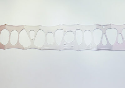 A long, horizontally stretched band of fabric in gradient shades from brown to white to pink, with oval cutouts, is mounted on a white wall. This artwork showcases the Art of ReUse through its creative use of repurposed materials.