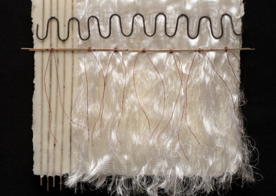 A mixed-media textile artwork with wavy metal, wooden rods, white ribbed and shiny fabric, and long, silky white threads hanging down—all arranged on a black background—celebrates the Art of ReUse in striking sustainable art form.