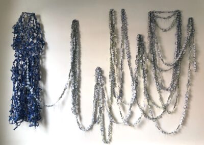 Crocheted blue and white chains hang artfully on a white wall, showcasing Upcycled Art with the left section denser and the right side forming loose, cascading loops.