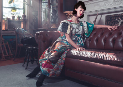 A woman in a colorful, patterned dress sits on a brown leather sofa in a vintage, sunlit room filled with plants, art, and ornate decor. Celebrating the Art of ReUse, she leans gracefully, her eyes closed and face partially in shadow.