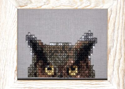 Cross-stitch art by Shawn Holtaway in a light wooden frame shows the top half of an owl’s head with yellow eyes and pointed ears against a white background, perfect for adding a touch of Keller style to your space.