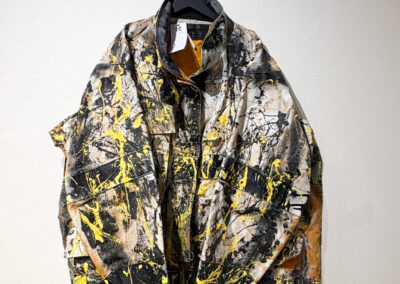 A jacket with a bold abstract design hangs on a black hanger against a white wall. The fabric features black, yellow, and white paint splatters and brush strokes, giving it an artistic, graffiti-like appearance.