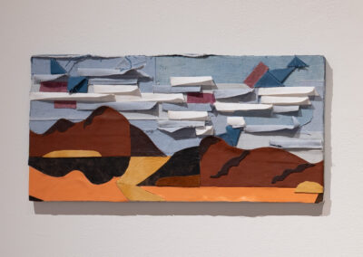 A textured artwork depicting a landscape with brown mountains, blue sky, and white clouds made from layered, cut pieces of fabric or paper, mounted on a plain white wall.