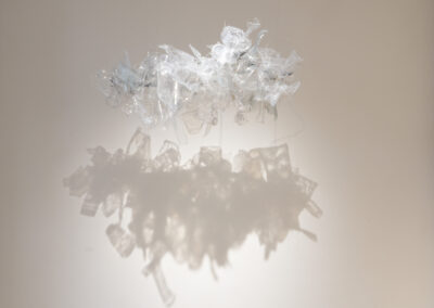 A cluster of translucent, crumpled plastic pieces hangs suspended against a plain wall, casting a soft, intricate shadow below on the surface illuminated by gentle lighting.