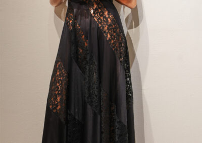 A mannequin with a bronze finish is dressed in a long, sleeveless black dress featuring sheer lace panels and a V-neckline, standing against a plain light-colored wall and wooden floor.