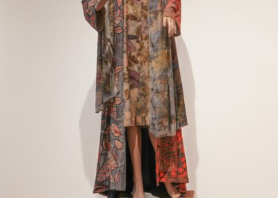 A mannequin wearing a patterned hat, a multicolored knee-length dress, and a long patterned coat stands on a small wooden stool against a plain white wall.