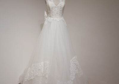 A white lace wedding dress with a fitted bodice, sleeveless design, and a full tulle skirt featuring lace detailing, displayed on a dress form against a plain wall and wooden floor.