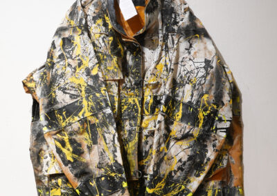 A jacket with an abstract design featuring black, white, and yellow paint splatters hangs on a black hanger against a white wall.