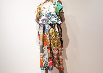 A mannequin dressed in a colorful, patchwork outfit featuring a ruffled short-sleeve top and wide-leg cropped pants, standing against a plain white wall and light wood floor.