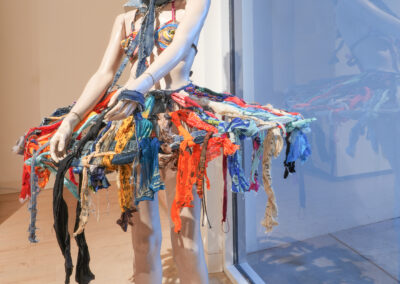 A mannequin wearing a knitted mask and a colorful, fringed dress made of various fabric strips stands by a large window in a brightly lit gallery space.