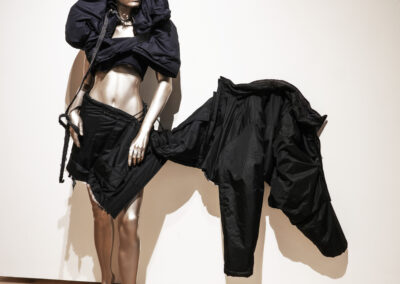 A silver mannequin stands against a plain wall, wearing an avant-garde black outfit that dramatically extends to the side, forming sculptural shapes with voluminous and structured fabric.
