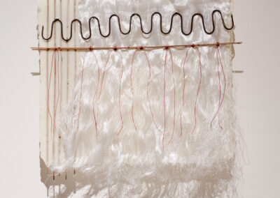 Textile artwork featuring a wavy black wire attached to a rod, from which long, white, silky threads hang down. The piece is mounted on a textured surface with vertical lines on the left side.