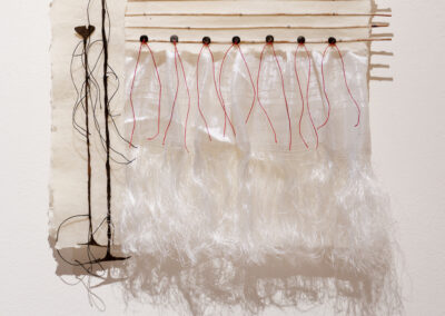 A mixed-media artwork features delicate, translucent white fabric strips hanging from wooden rods, with red threads and organic, root-like forms extending down the left side, all mounted on textured white paper.