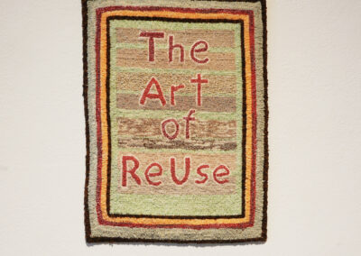 A handmade textile hanging on a white wall displays the words "The Art of ReUse" in large, red letters, surrounded by multicolored striped borders.