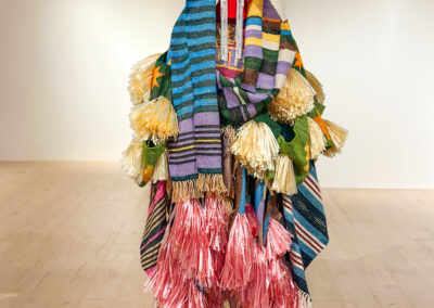 A person stands covered head to toe in a colorful costume made of layered striped fabrics, gold and pink tassels, and fringes, with only their feet and a hint of their face barely visible. The background is a plain gallery space.
