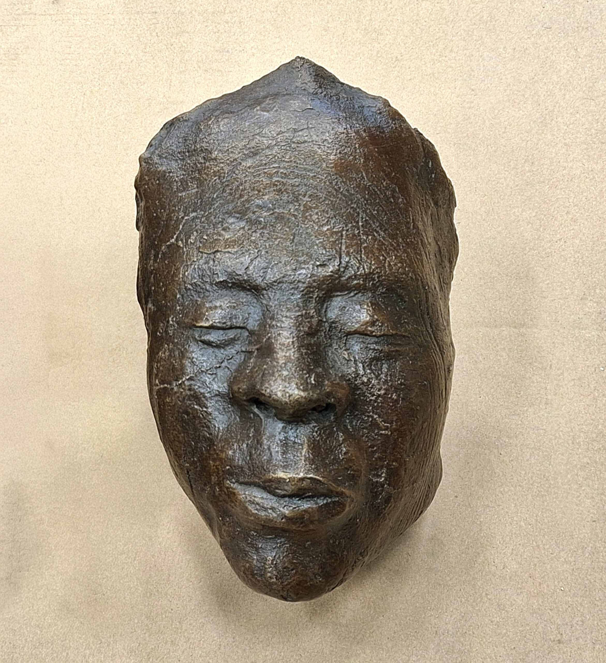 A dark, textured sculpted mask of a human face with closed eyes and relaxed expression, displayed against a plain beige background.