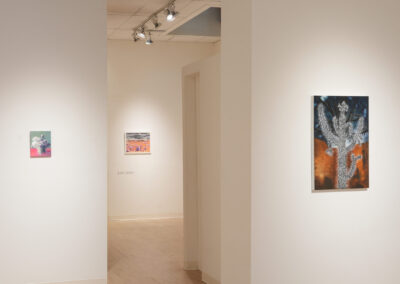 A minimalist art gallery with white walls displays three colorful, abstract artworks. Track lights illuminate the pieces, and a narrow hallway leads to another room. The space is clean and modern.