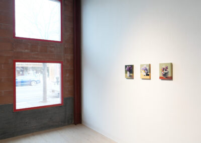A minimalist gallery space with two large windows on the left and three small colorful still-life paintings of flowers hanging on a white wall to the right. Natural light fills the room.