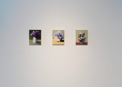 Three small paintings of floral still lifes are evenly spaced and mounted on a plain white wall. Each painting features a vase with flowers, using soft, muted colors on simple backgrounds.