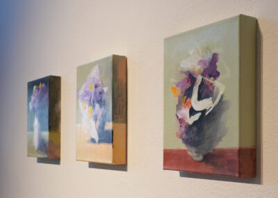 Three abstract floral paintings on canvas hang on a cream-colored wall, each with loose, colorful brushstrokes depicting vases of flowers against muted backgrounds.