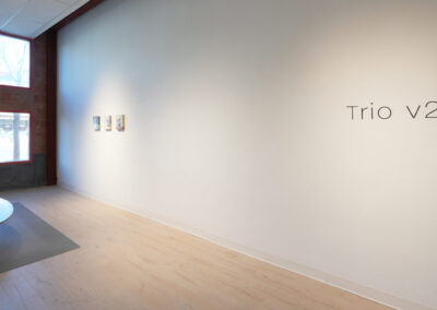 Minimalist gallery space with light wood floors, a round table on the left, and three small artworks on a white wall. The text "Trio v2" is displayed on the right side of the wall. Large windows let in natural light.
