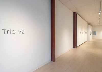 A minimalist art gallery with light wood floors and white walls, featuring the text "Trio v2" on the left wall and small artworks displayed along the right wall. The space is brightly lit with ceiling spotlights.