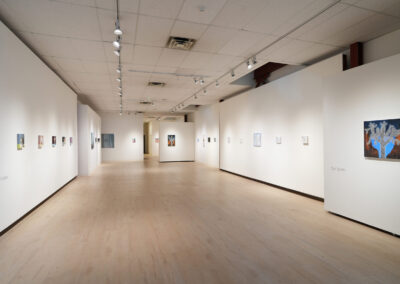 A spacious, well-lit art gallery with light wood floors and white walls, displaying small framed artworks evenly spaced along both sides. Track lighting illuminates the artwork, creating a calm, modern atmosphere.