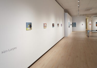 A contemporary art gallery with small paintings displayed along white walls and "Kim Largey" written on the left wall. The space has light wood floors and ceiling spotlights.