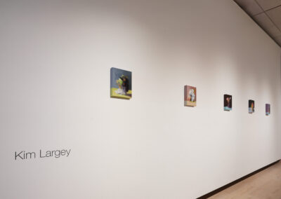 A white gallery wall displays six small colorful paintings in a row. The artist's name, "Kim Largey," is written on the left side of the wall in black text. The floor is light wood.