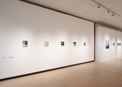A gallery with light wood floors and white walls displays small framed artworks in a row. "Kim Largey" is written on the left wall. Ceiling spotlights illuminate the paintings.