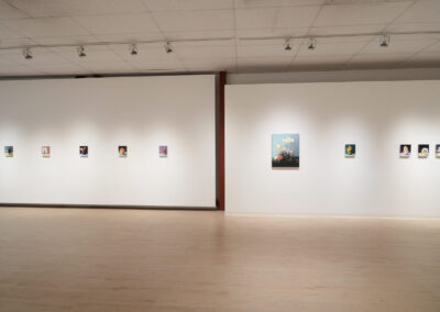 An art gallery with white walls displays small framed artworks in a row, spaced evenly apart. The room has light wood floors, a tiled ceiling, and track lighting illuminating the pieces.