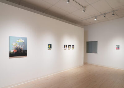 A modern art gallery with white walls displays several framed paintings, including still lifes and portraits, spaced evenly along the wall. Track lighting illuminates the artwork. The room has light wood floors and a window on the right.