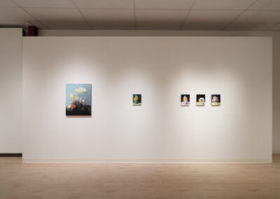 A white gallery wall displays five paintings: one large floral still life on the left and four smaller colorful portraits in a row to the right, all under soft overhead lighting on a light wood floor.