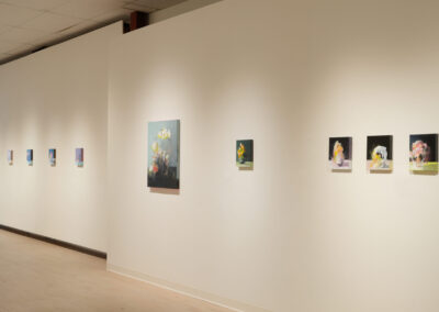 A gallery wall displays a series of small, colorful floral paintings in a row, with one larger painting centered. The room has light wood floors and neutral-colored walls.