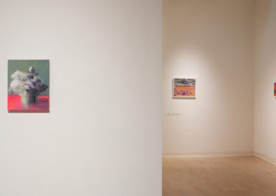 A gallery with three paintings on white walls; one abstract piece with a figure on the left wall, and two colorful landscapes on the far wall. The room has wooden floors and minimal lighting.