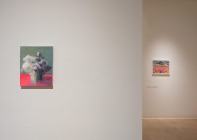 A gallery wall displays a painting of white flowers in a vase on a bright pink surface. In the background, another artwork and the name "Julie Green" are visible on a separate wall. The space is minimal and well-lit.