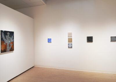 A gallery wall displays six small abstract artworks in a row, featuring various colors and textures, with a larger painting of a cactus on the left; wooden floor and white walls complete the minimalist setting.