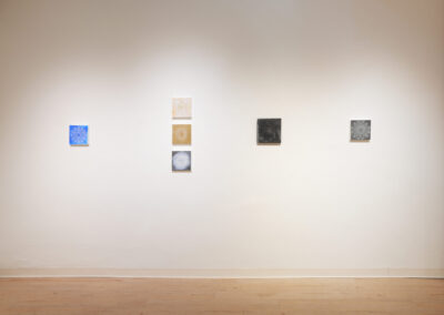 A minimalist art gallery wall displays seven small square artworks arranged in two vertical columns with spacing between them, featuring various abstract designs in blue, beige, black, and gray tones. The floor is light wood.