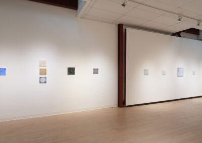 A gallery space with white walls displays small, square artworks arranged in single rows. The pieces vary in color and design, and the room has wooden floors and a white ceiling with spotlights.