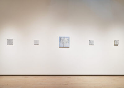 A gallery wall displays five small square artworks in a row; the central piece features a faint blue tone, while the other four appear monochromatic. The wall is white and the floor is light wood.