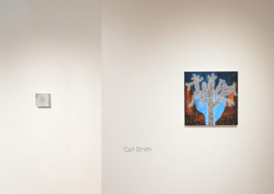 Two artworks hang on a white gallery wall. The left piece is a small, square, monochrome work. The right is a larger, colorful painting of a blue tree with white dots. The name "Carl Smith" is written below them.